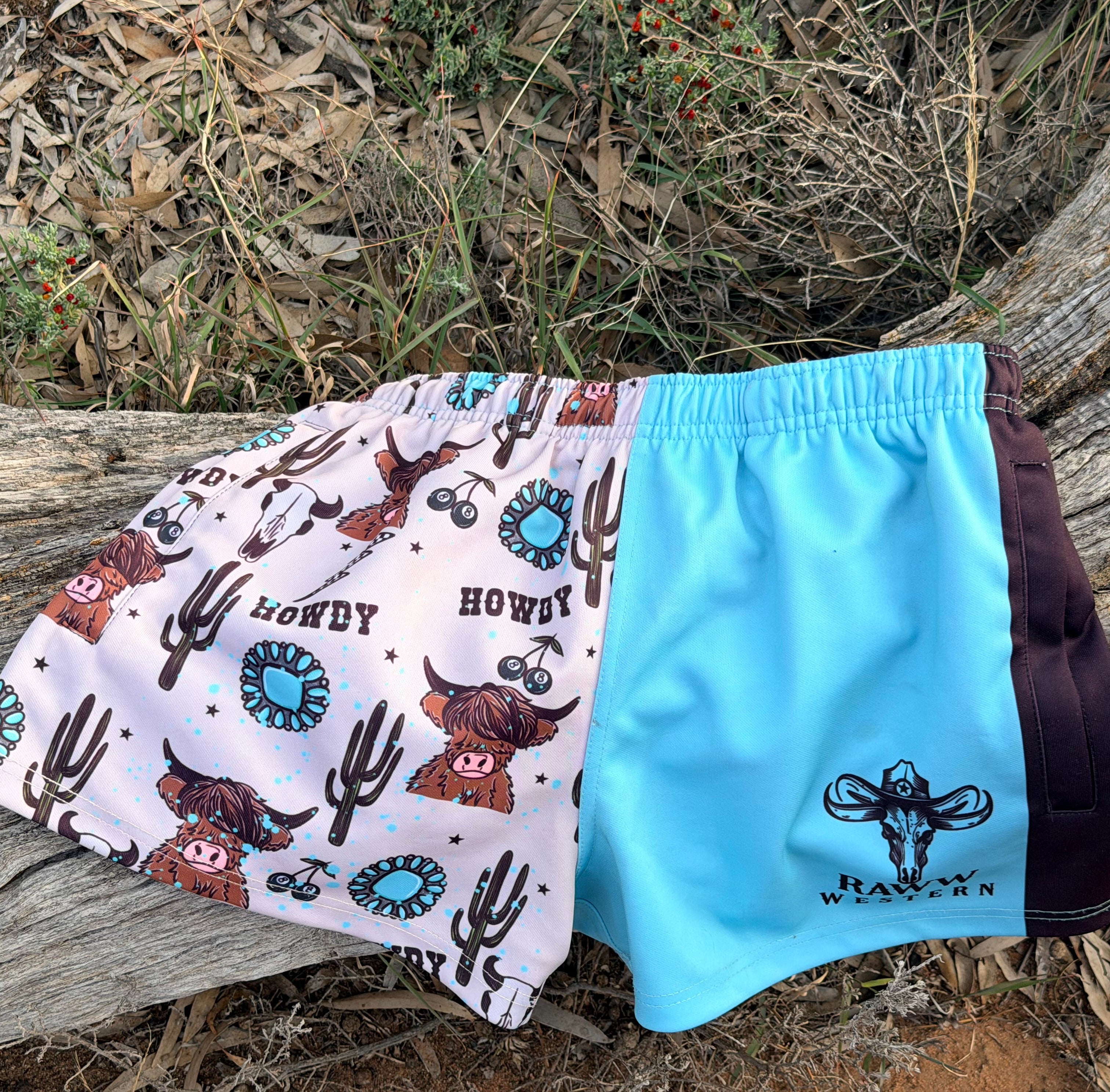 Footy shorts laid flat: white cow print fabric with blue drawstring and RAW WESTERN